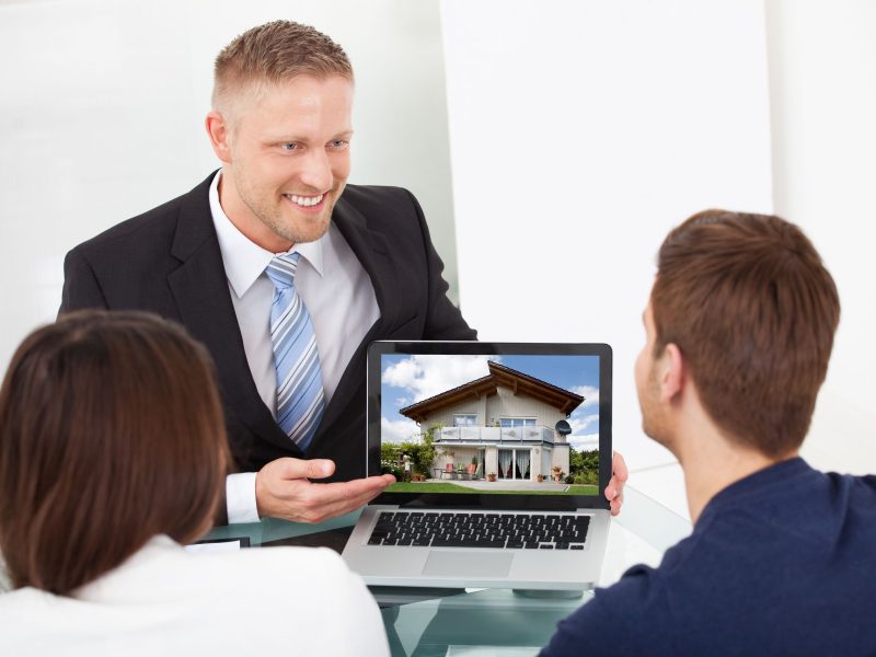 28161926 - smiling advisor showing house picture to couple on laptop at office desk